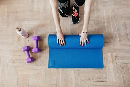 Iemand rolt een blauwe yogamat uit op een houten vloer, naast twee paarse dumbbells en een bidon
