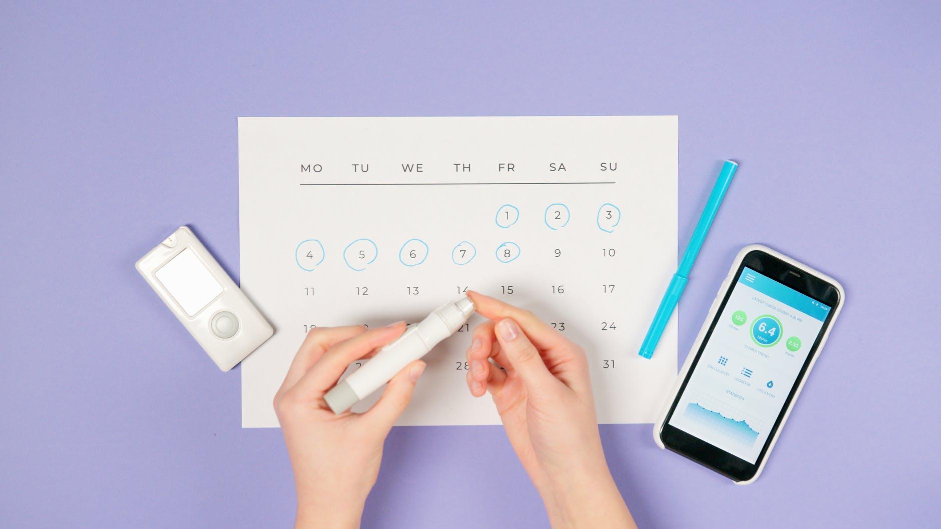 Handen met een insulinepen boven een kalender, naast een glucosemeter en een telefoon met een diabetes-app