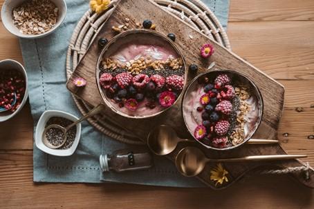 Twee yoghurtbowls met frambozen, bosbessen, granola en chiazaad op een houten plank